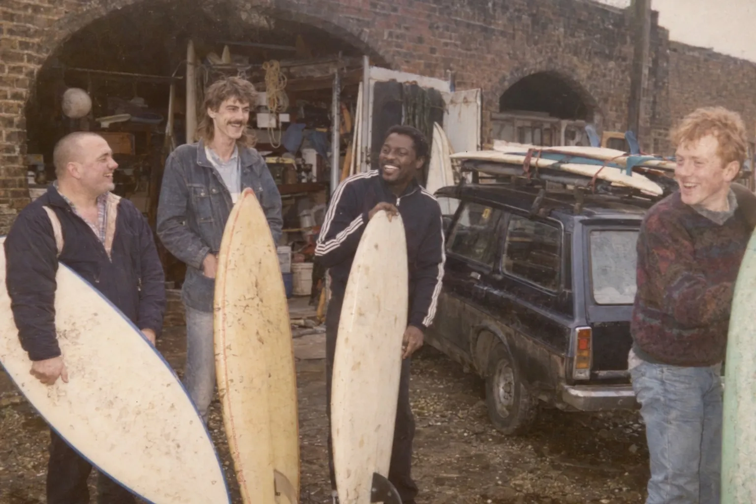 A faded 1980s photo of four young men holding surfboards outside a railway arch, an estate car with boards on the roof beside them
