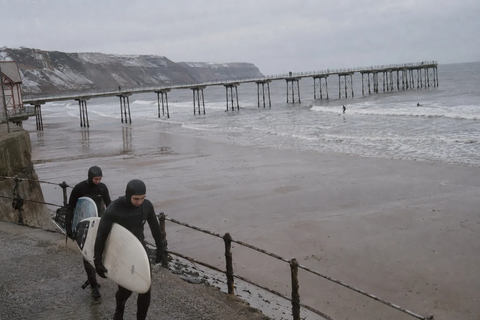 Two surfers walk down the slipway beside Saltburn pier on an overcast morning