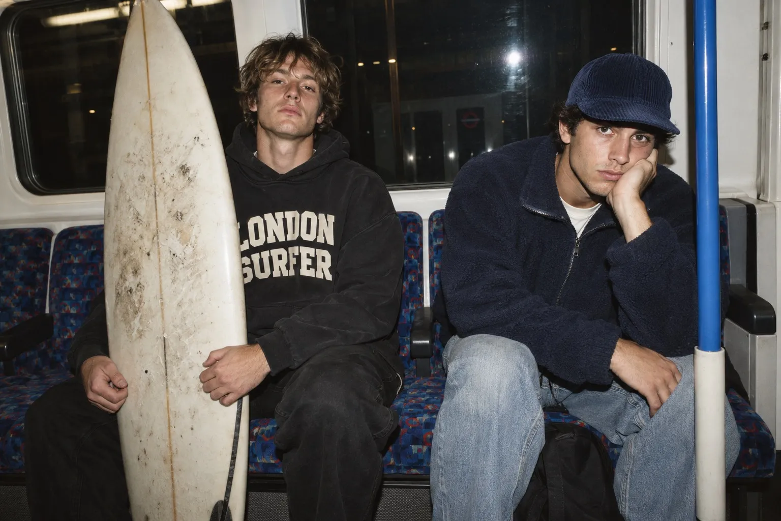 Two friends on a late-night Tube with a surfboard